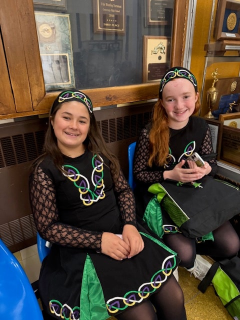 Two young dancers in Celtic costumes seated in front of a trophy case