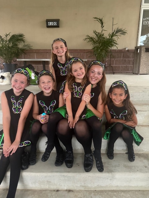 Five young dancers in Celtic costumes smiling together under an outdoor pavilion