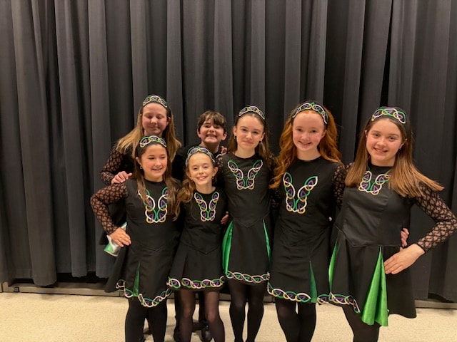 Group of seven young dancers in black and green Celtic Irish dance costumes posing indoors
