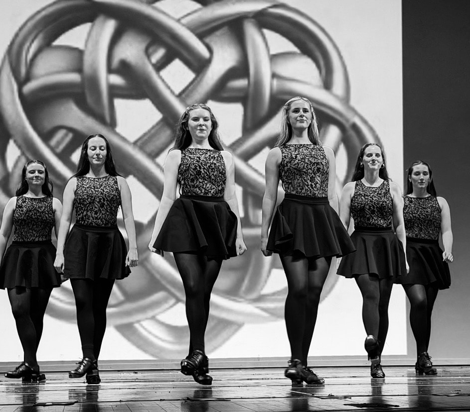 Six adult dancers in black lace costumes performing in front of a Celtic knot backdrop, black and white photo