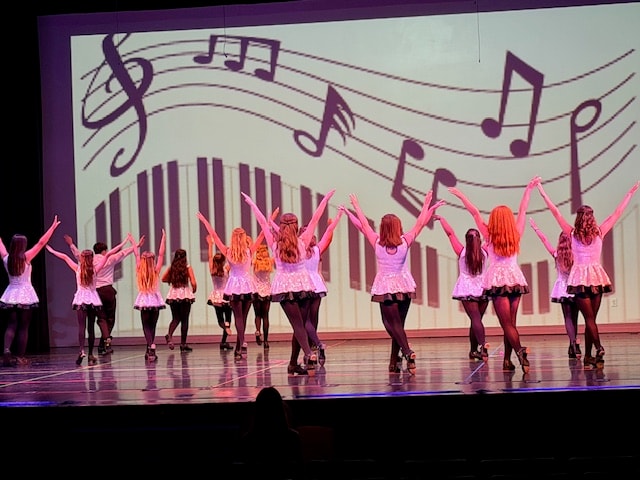 Dancers with arms raised on stage in front of a musical notes backdrop