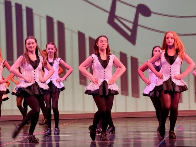 Dancers in silver vests and black skirts performing with musical notes backdrop