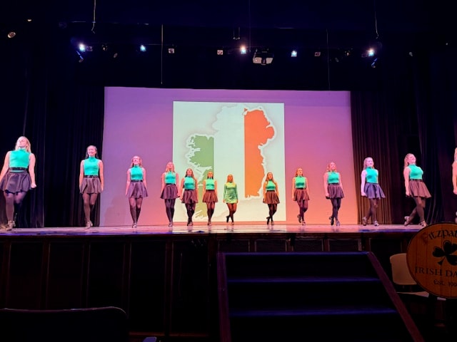 Full group of Fitzpatrick dancers in teal tops on stage with Ireland map backdrop